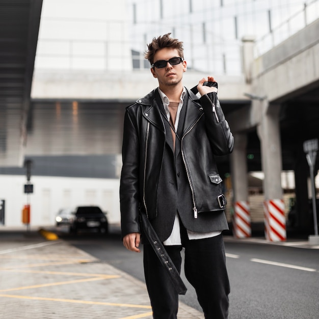 American fashion young man model in a stylish oversized leather black jacket in dark sunglasses in vintage jeans walks on the street in the city.Trendy handsome hipster guy stands near road outdoors.