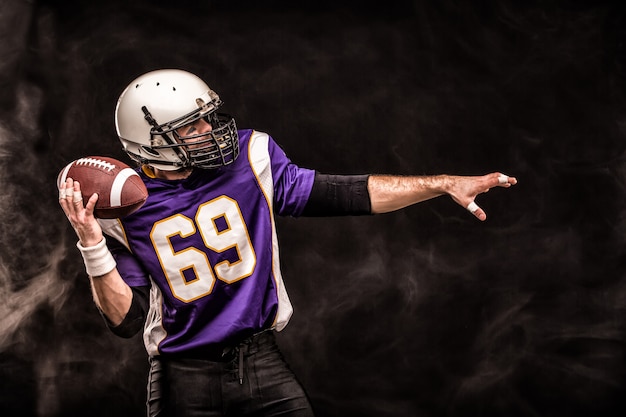 American football player holding ball in his hands in smoke. Black background, copy space.