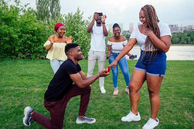 american male standing on one knee asking woman marriage giving ring proposing surprised shocked while gathering group friends all around in shock confusing evening cloudy autumn spring in the park