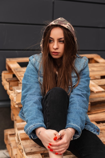 American modern pretty stylish young woman in youth fashionable denim clothes with a military hood posing on wooden pallets outdoors. Urban trendy girl fashion model resting in the city. Street style.