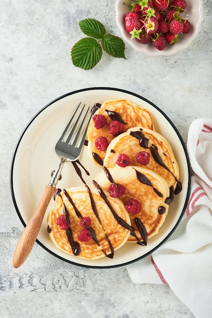 American pancakes Pancakes with fresh raspberry with chocolate glaze or toppings in gray bowl on light gray table background Homemade classic american pancakes Page for magazine concept Top view