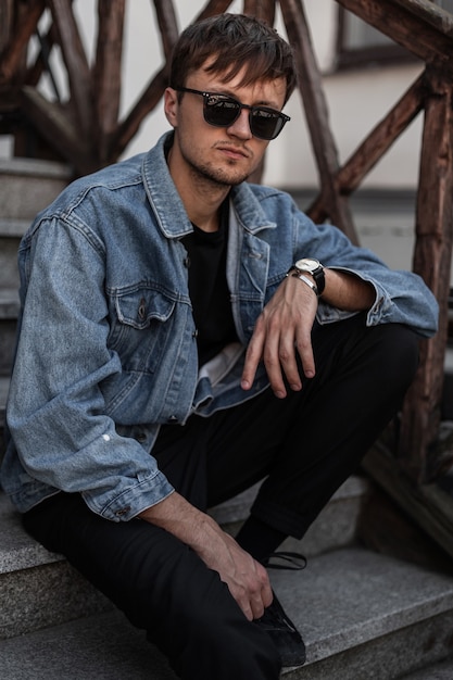 American young man in a blue denim jacket in trendy pants in stylish black sunglasses relax sitting on a vintage staircase in the street on a spring day. Fashionable guy hipster enjoys a rest outdoors