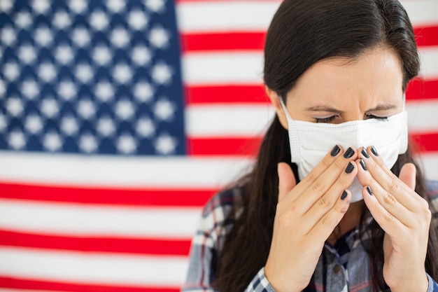 American young woman with the USA flag in the background . Wearing a mask and with coronavirus symptoms