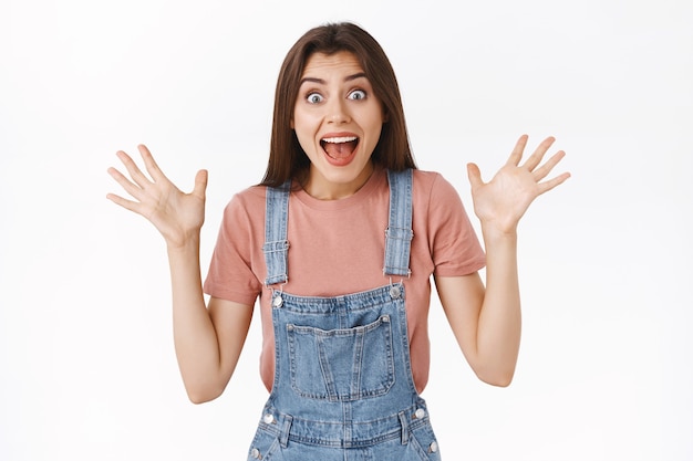 Amused, excited cheerful beautiful brunette woman in overalls, t-shirt, raising hands up thrilled and enthusiastic, scream from amazement, triumphing, look surprised as win lottery