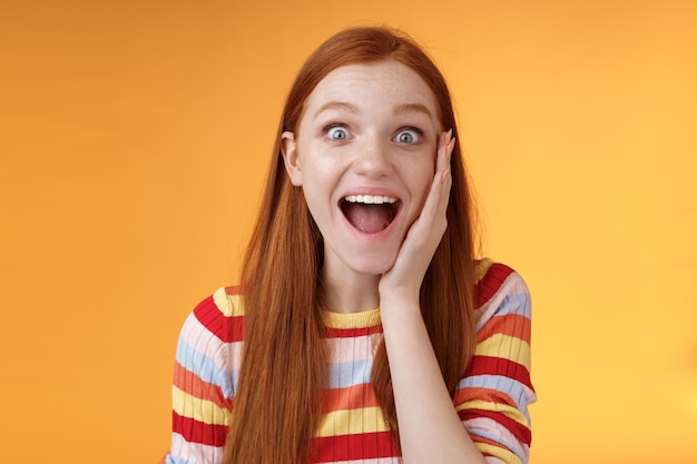 Amused happy smiling good-looking redhead woman screaming happily touch cheek astonished receive good perfect news triumphing feeling excited look thrilled camera, standing orange background.