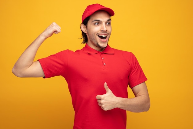 amused young delivery man wearing uniform and cap looking at side showing strong gesture and thumb up isolated on yellow background