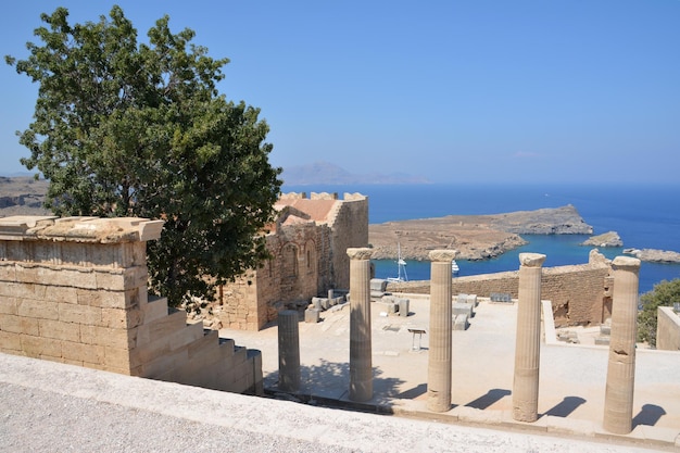 ancient columns of acropolis of greek town Lindos with green tree and lagoon on background