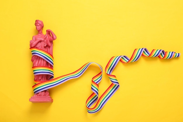 Ancient Greek goddess statue with LGBT rainbow ribbon pride tape symbol on yellow background Top view