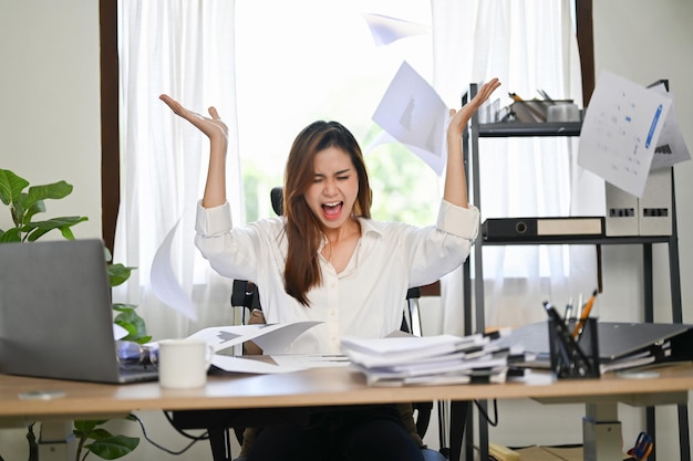 Angered and mad Asian businesswoman screaming and throwing up papers at her desk