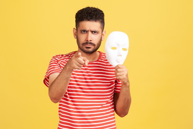 Angry annoyed man with beard in striped t-shirt pointing finger on you holding white mask with unknown face, blaming you in duplicity. Indoor studio shot isolated on yellow background