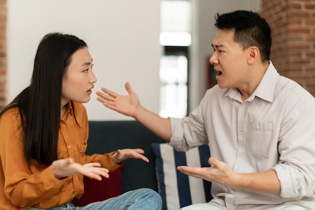 Angry asian spouses having quarrel experiencing crisis in relationship sitting together on sofa at home