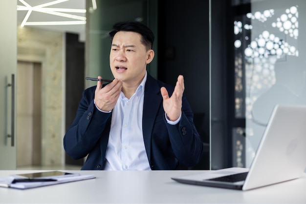 Angry asian young man businessman emotionally talking on the phone through loudspeaker solving