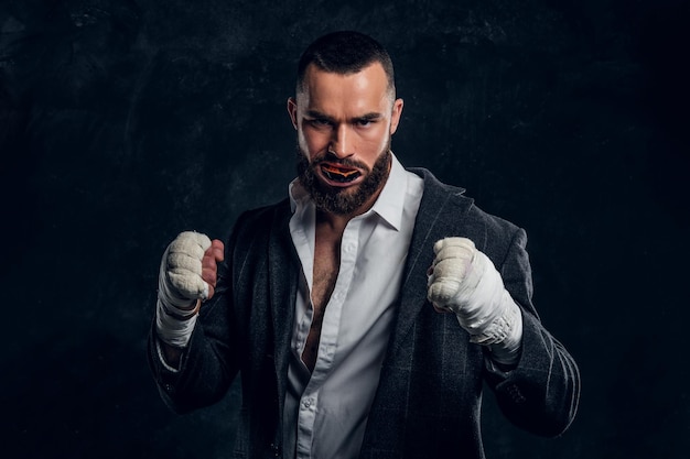 Angry bearded businessman in suit and protective kickboxing gloves is posing for photographer at dark photo studio.