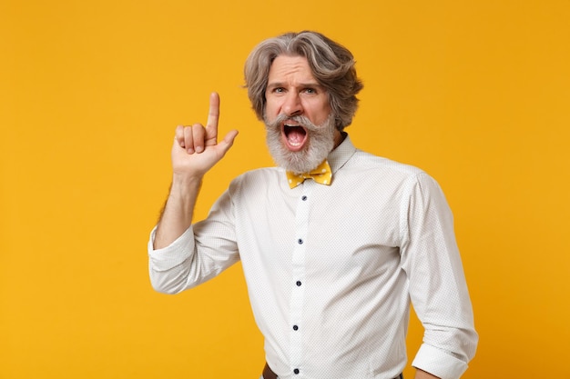 Angry elderly gray-haired mustache bearded man in white shirt bow tie posing isolated on yellow background. People lifestyle concept. Mock up copy space. Screaming, swearing, pointing index finger up.