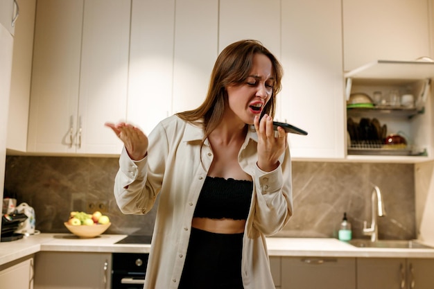 Angry emotional woman shouting expressively to smart phone while answering phone call or recording a voice message while cooking at modern kitchen