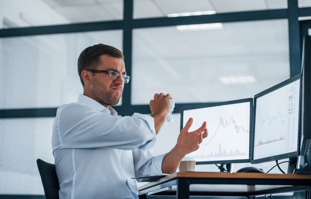 Angry employee throws paper. Conception of failure. Young businessman in formal clothes is in office with multiple screens.