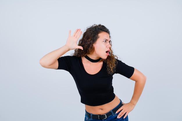 Angry girl is holding hand near ear and putting other hand on waist on white background