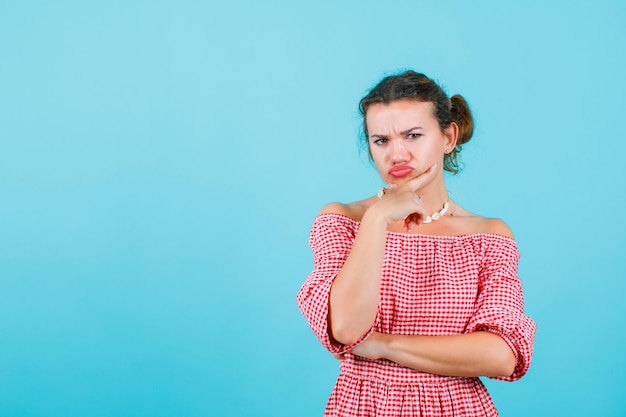 Angry girl is looking at camera by holding hand on chin on blue background