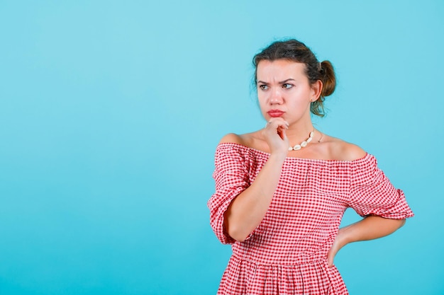 Angry girl is thinking by holding one hand on chin and other on waist on blue background