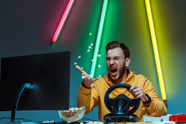 Angry and goodlooking man in glasses playing with steering wheel and throwing popcorn