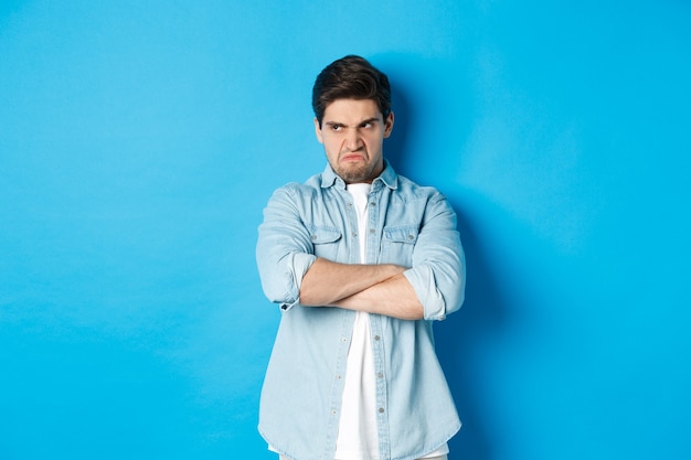 Angry guy cross arms on chest and looking away with insulted expression, standing offended against blue wall