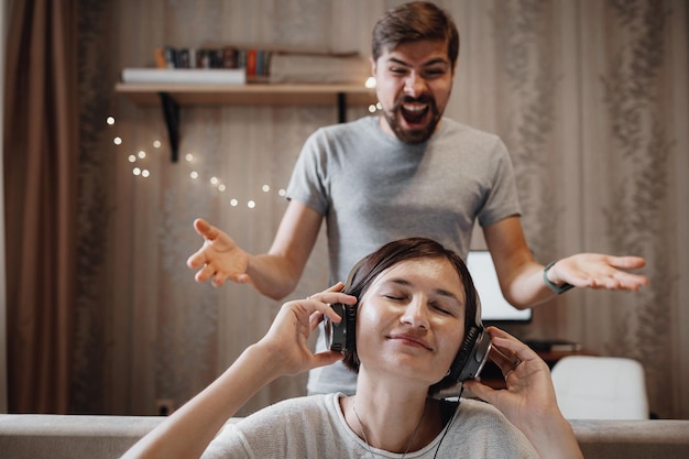 Angry husband shouting at wife sitting on sofa and covering ears with headphones