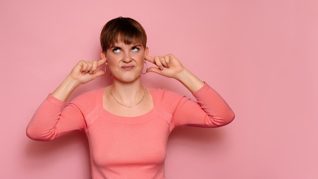 An angry irritated young woman isolated on a pink background covers her ears from loud noisesHuman facial expressions emotions Body language Copying space