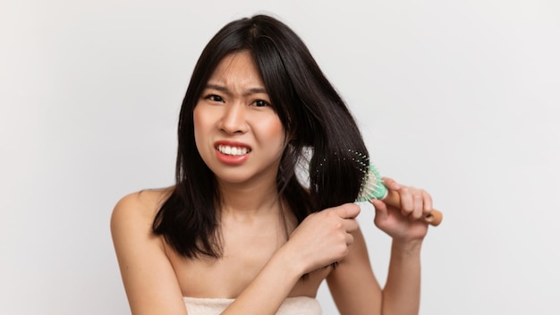 Angry japanese lady having problem of thick hair while using hair brush combing hair over white