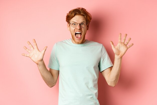 Angry and pressured young man losing temper, spread hands sideways and screaming with furious face, standing in glasses and t-shirt against pink background