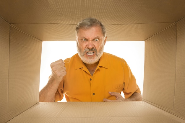 Angry senior man opening the biggest postal package isolated on white. Angry male model on top of cardboard box