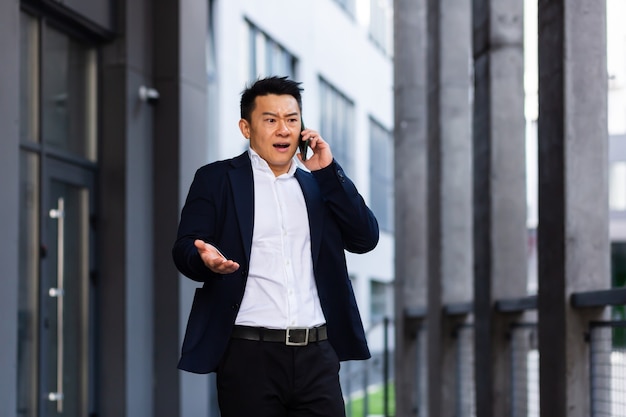 Angry and serious Successful Asian businessman explains information to employees using phone, speaks near office outside