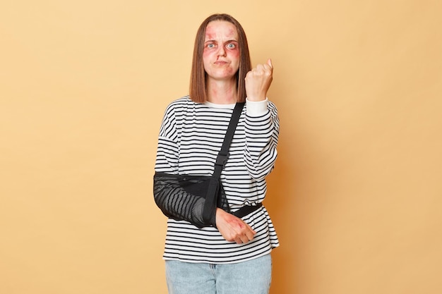 Angry sick woman with scratches and bruises on her face standing isolated over beige background showing fist after being beating revenge