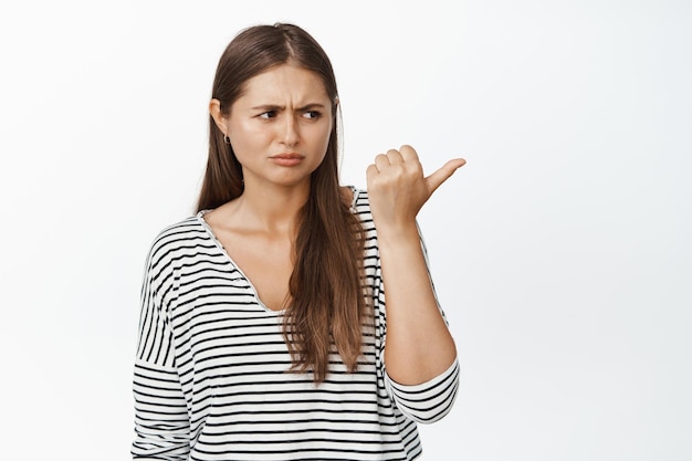 Angry sulking woman pointing finger right, looking at smth unfair, showing upset expression, standing in casual clothes against white background.