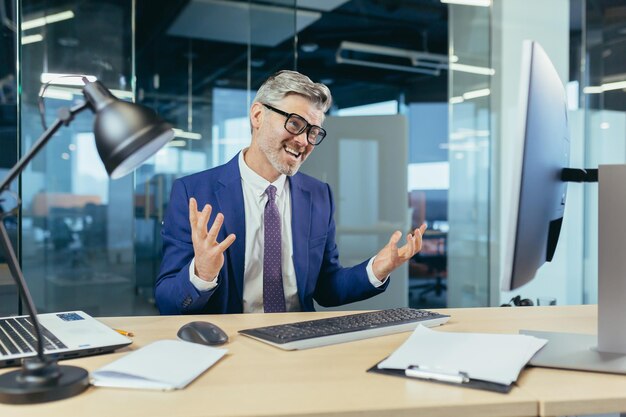 Angry and upset boss businessman shouting at computer monitor man with glasses working in modern office at computer