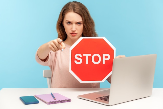 Angry woman boss office employee at workplace holding stop symbol and pointing to camera showing red traffic sign negative prohibition concept indoor studio shot isolated on blue background