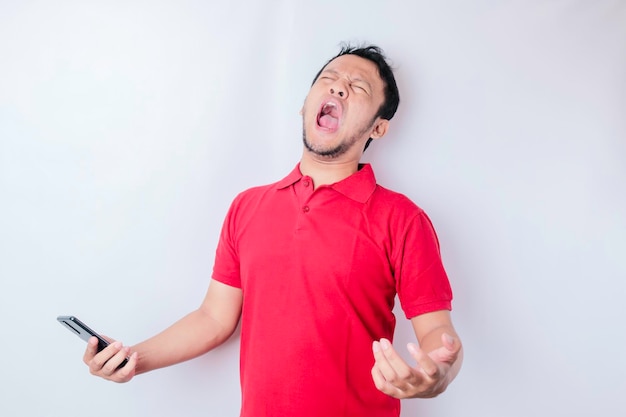 An angry young Asian man looks disgruntled wearing red shirt irritated face expressions holding his phone