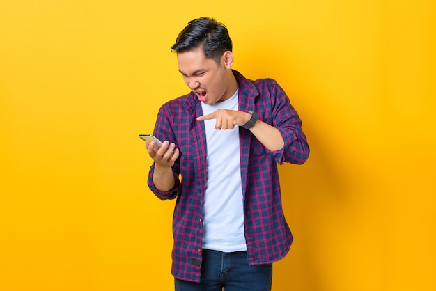 Angry young Asian man in plaid shirt using a mobile phone and yelling isolated on yellow background