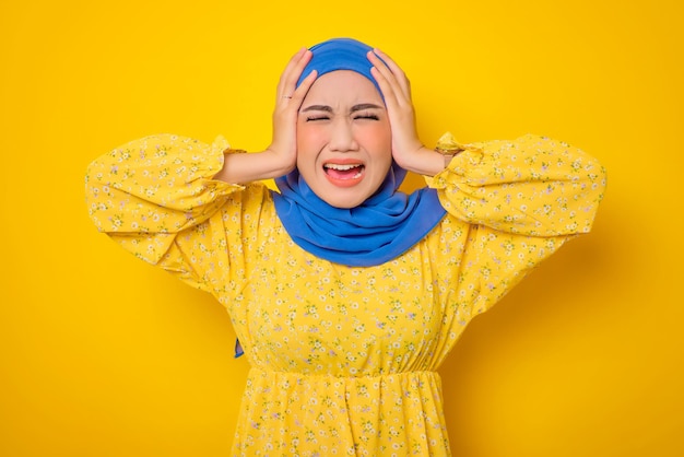 Angry young Asian woman in casual dress screaming with covering her ears isolated on yellow background