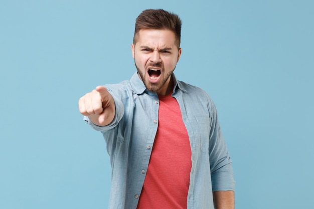 Angry young bearded guy in casual shirt posing isolated on pastel blue background studio portrait. People lifestyle concept. Mock up copy space. Pointing index finger on camera, screaming swearing.