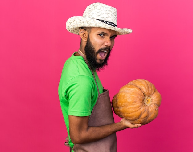 Angry young gardener afro-american guy wearing gardening hat holding pumpkin 