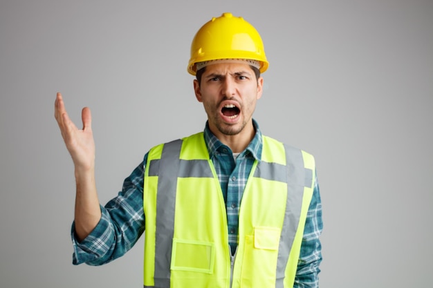 Angry young male engineer wearing safety helmet and uniform looking at camera keeping hand in air shouting isolated on white background