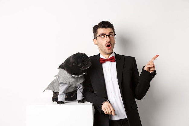 Animals, party and celebration concept. Amazed young man and black dog in costumes staring right at copy space, standing against white background