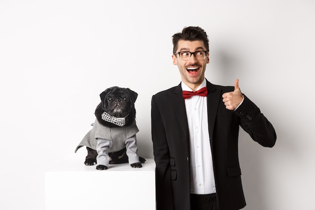 Animals, party and celebration concept. Handsome young man in suit and cute black pug in costume staring at camera, owner showing thumb up in approval and praise, white background