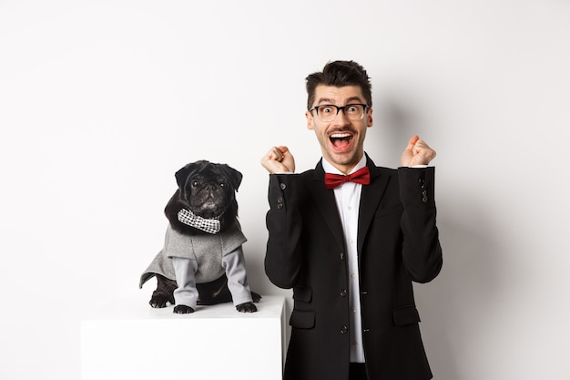 Animals, party and celebration concept. Happy young man in suit and puppy in pet cosume standing over white background, dog owner rejoicing and triumphing.