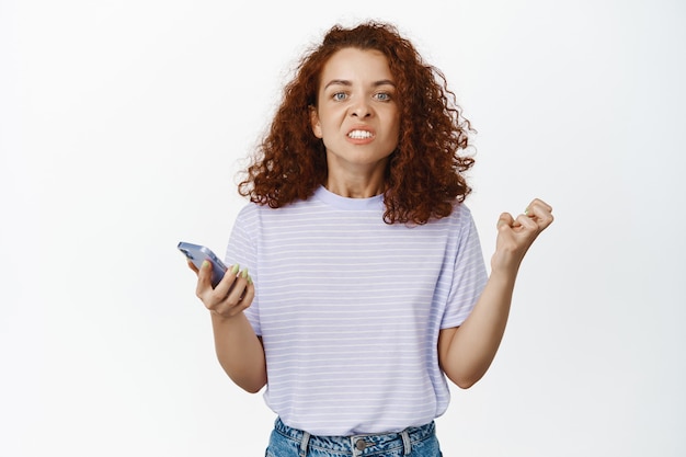 Annoyed and angry redhead woman looks pissed off, holds mobile phone and clenched fist in anger, irritated with message notification, bad UI UX design on white.