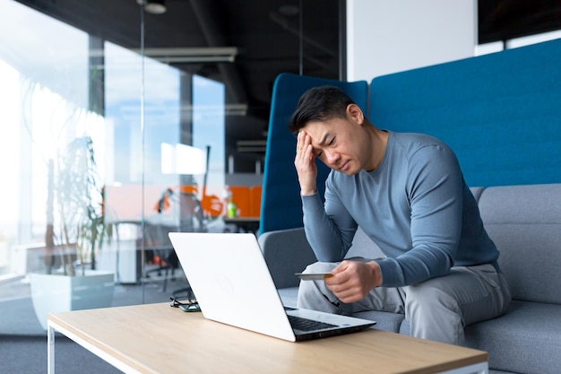 Annoyed asian man trying to do banking with laptop and credit card businessman in working in modern office