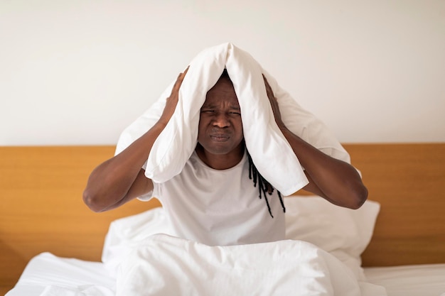 Annoyed Black Guy Covering Ears With Pillow While Sitting In Bed