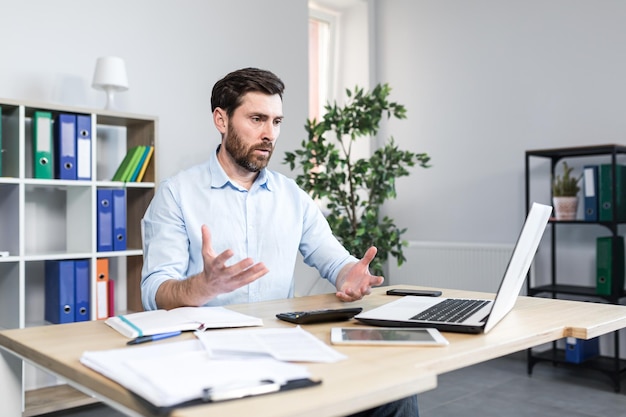 Annoyed emotional nervous young man office worker manager talking on a video call at the computer sitting at a table in the office in white clothes