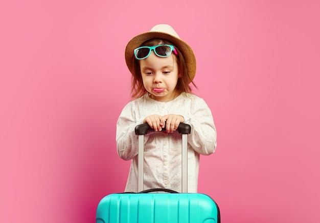 Annoyed and sad little girl with suitcase stands on pink isolated. Sadness