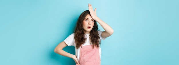 Annoyed and tired young woman in summer outfit slap forehead and roll eyes standing over blue backgr